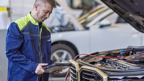 Ein Arbeiter steht mit Lachen neben Auto mit der Motorhaube offen und hat Tablet im Hochformat