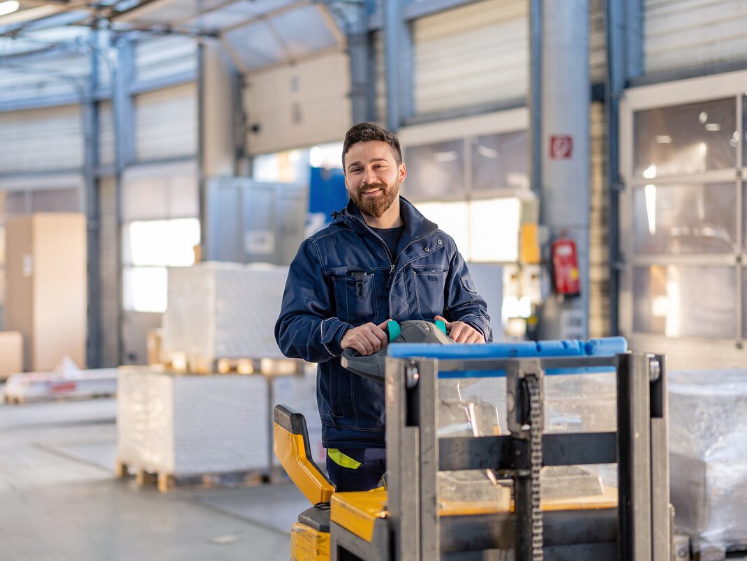 Ein Arbeiter lacht und faehrt mit einem beladenen Gabelstapler