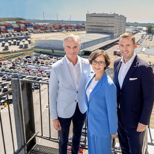 Gruppenbild - Dame mit zwei Herren auf Balkon mit Fahrzeugparkplatz im Hintergrund