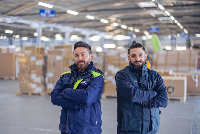 Es stehen zwei Mitarbeiter mit gekreuzten Armen in einer Lagerhalle
