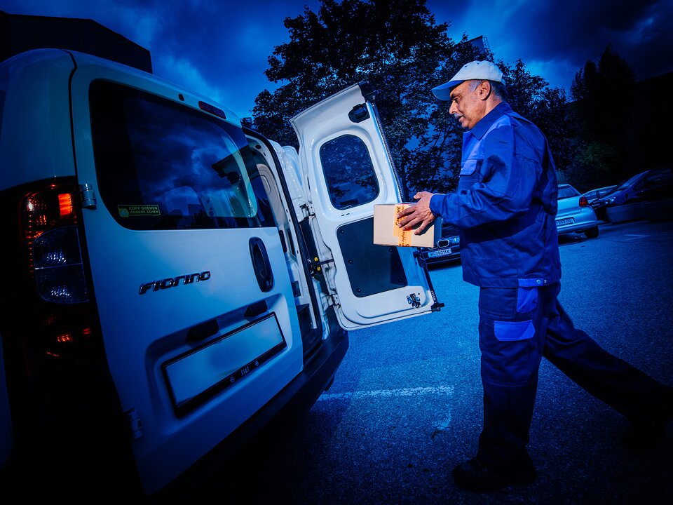 Mann bringt Paket zu offener Hecktür bei Fahrzeug.