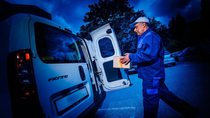 Mann bringt Paket zu offener Hecktür bei Fahrzeug.