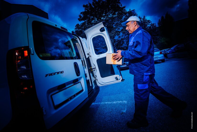 Mann bringt Paket zu offener Hecktür bei Fahrzeug.