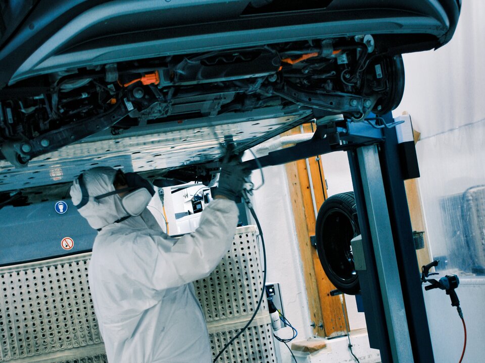 Fahrzeug auf Hebebühne Mann mit Maske arbeitet an Unterbodenkonservierung Querformat Lagermax
