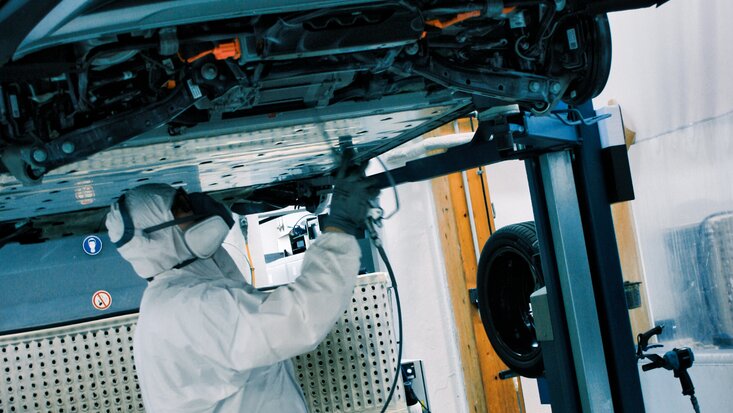 Fahrzeug auf Hebebühne Mann mit Maske arbeitet an Unterbodenkonservierung Querformat Lagermax