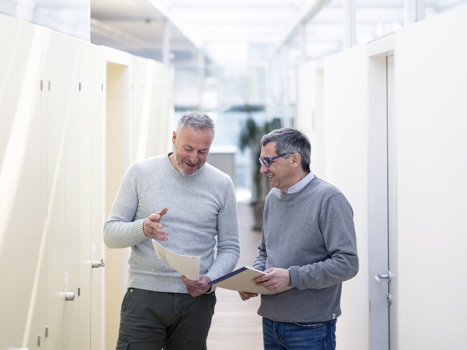 Zwei Mitarbeiter halten Unterlagen am Gang bei Besprechung in der Hand.