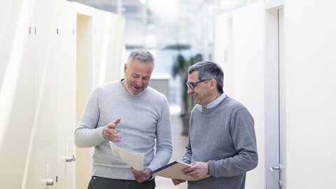 Zwei Mitarbeiter halten Unterlagen am Gang bei Besprechung in der Hand.