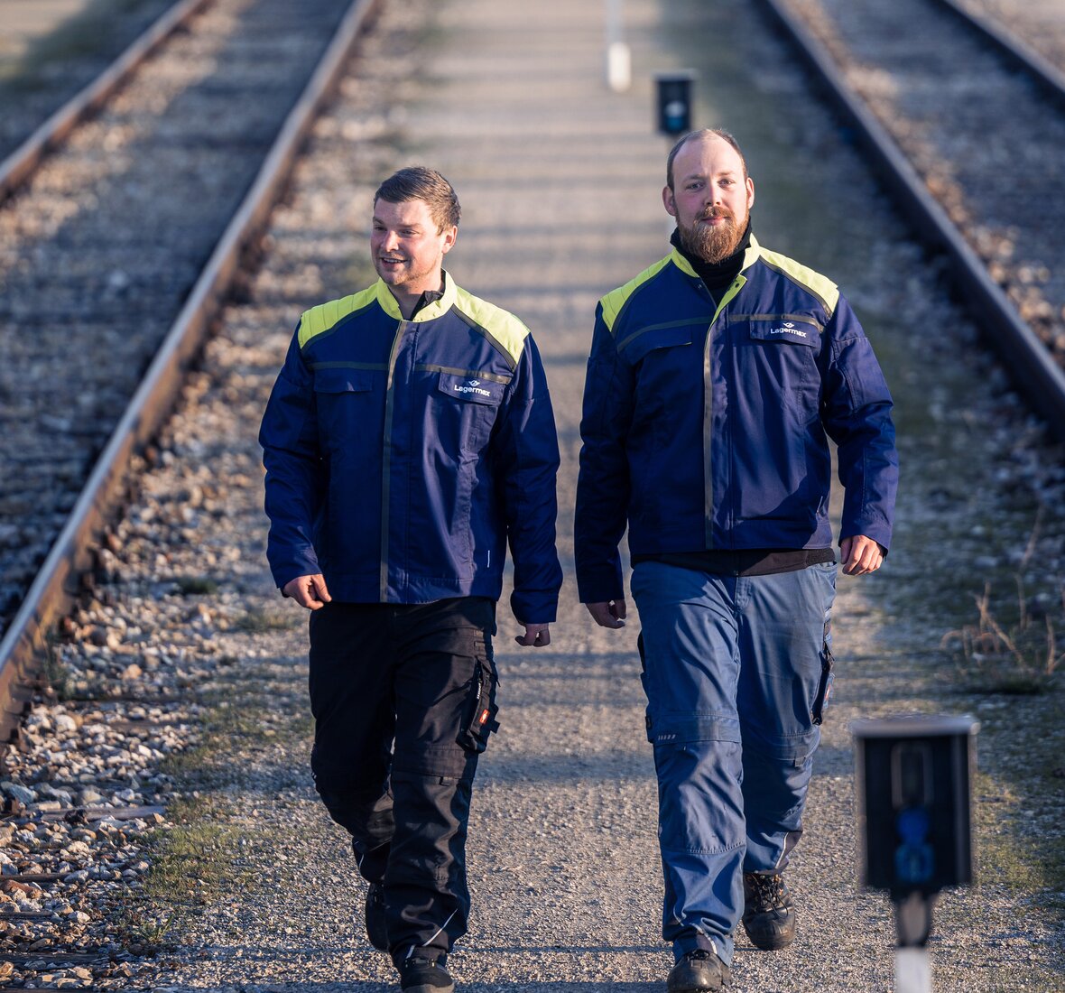 Zwei Lagermax Mitarbeiter in Arbeitskleidung gehen bei Bahnschienen