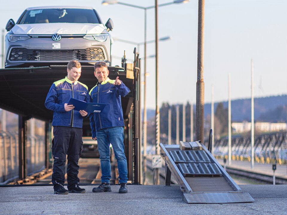 Zwei Lagermax Mitarbeiter stehen nebeneinander vor einem Autotransport-Lader mit einer Arbeitsmappe, und der rechte zeigt in die Ferne.