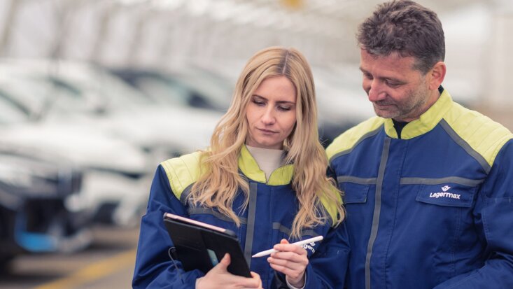 Frau und Mann stehen nebeneinander und arbeiten an einem Klemmbrett und im Hintergrund sieht man Autos gelagert.