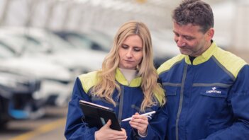 Frau und Mann stehen nebeneinander und arbeiten an einem Klemmbrett und im Hintergrund sieht man Autos gelagert.