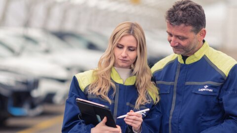 Frau und Mann stehen nebeneinander und arbeiten an einem Klemmbrett und im Hintergrund sieht man Autos gelagert.