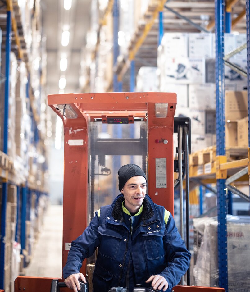 Man sieht einen Gabelstapler Fahrer Arbeiter in Nahaufnahme in einer Paketversand Halle mit Paketen links und rechts aufgebaut