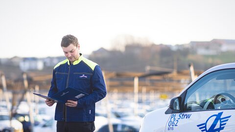 Lagermax Arbeiter hat Arbeitsmappe und blickt in diese fokussiert neben einem Arbeitsfahrzeug.