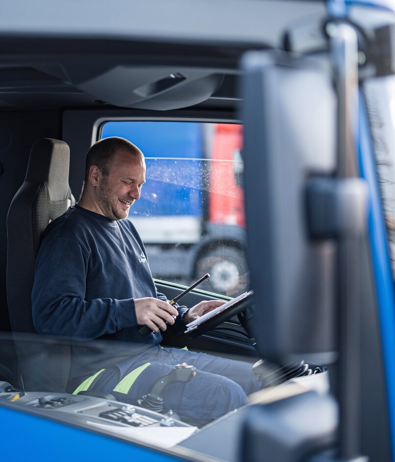 Ein Arbeiter sitzt in einer LKW Fahrerkabine und lacht mit Dokument und Funkgeraet in der Hand